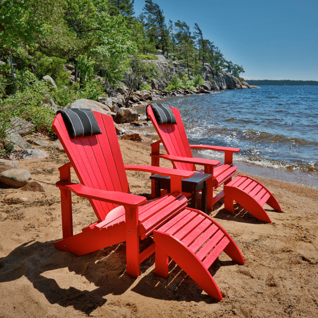Two red C.R. Plastics Adirondack chairs on a sandy beach by a lake